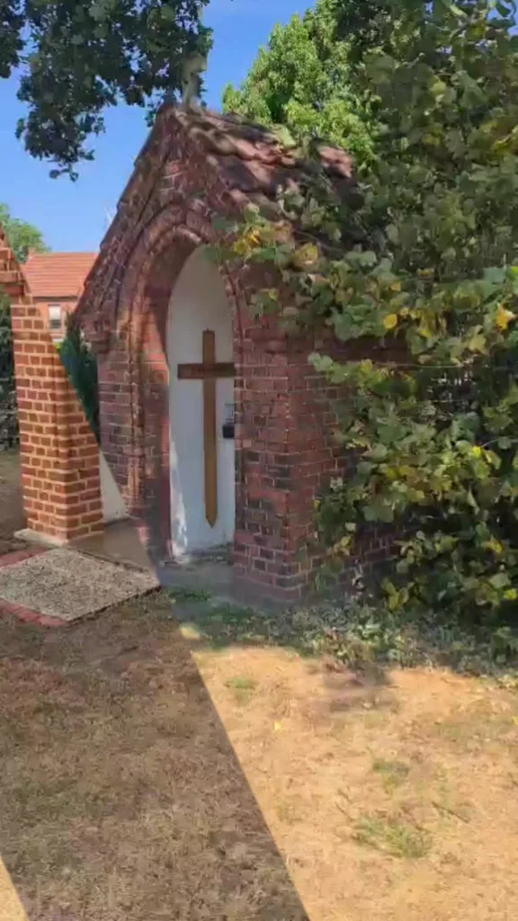 Eine kleine Backsteinkapelle mit weißer Tür und braunem Kreuz steht neben einem Laubbaum.