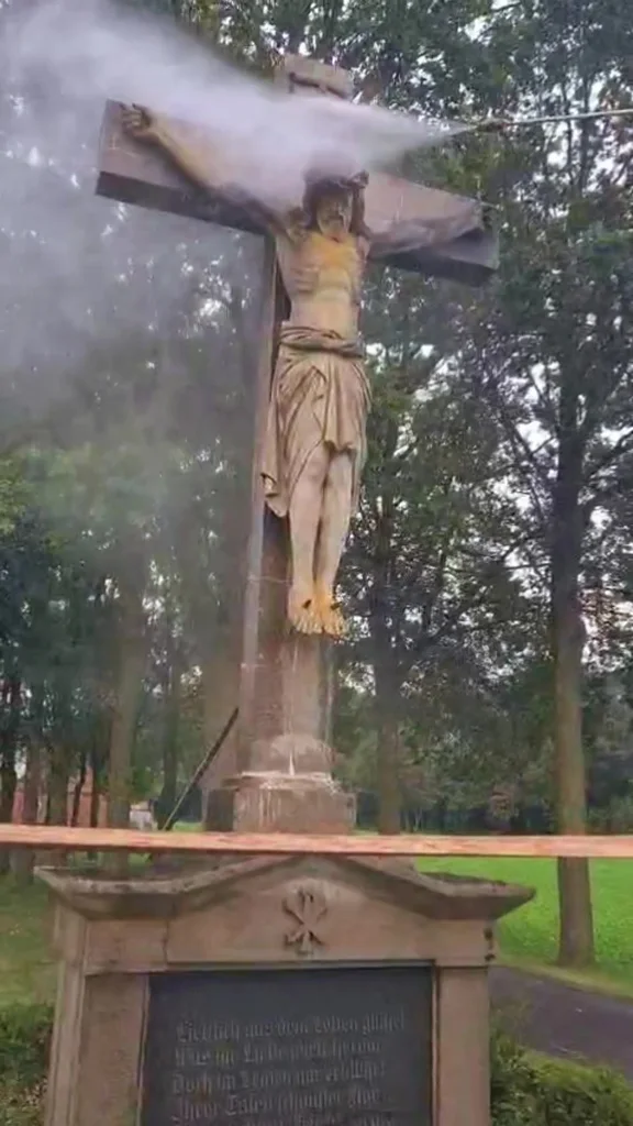 Ein Wasserstrahl trifft den oberen Teil eines steinernen Wegkreuzes mit Kruzifix unter Bäumen.